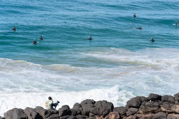 Coolangatta Avustralya - 4 Haziran 2024; kayalık kıyıların ötesindeki dalgalarda sörfçülerin küçük figürleri