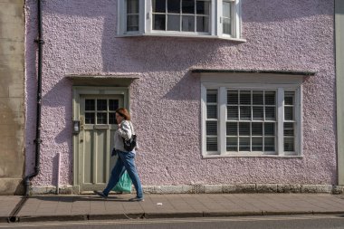 Oxford United Kingdom - July 24 2024; Woman waling in street scene passing old building with lilac wall and multi-paned windows. clipart