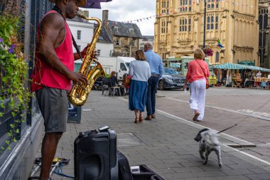 Cirencester Birleşik Krallık - 26 Temmuz 2024; Şehir sokak sahnesi kırmızı cüppeli sokak müzisyeni insanlar geçerken saksafon çalıyor.