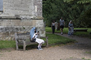 Stratford Upon Avon - Birleşik Krallık; 28 Temmuz 2024; Başının üzerinde mavi desenli peçe takan genç bir kadın güneş ışığından korunmak için kilise zemininde cep telefonu kullanıyor..