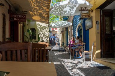 Chania Girit - 15 Ağustos 2024; Üç kişilik turist ailesi, kentin cennet dar sokaklarında ve caddelerinde gölgede serin içecekler içiyorlar..