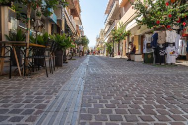Chania Girit - 15 Ağustos 2024; Bildiri tahtası, mavi sandalyeleri ve saksı bitkileriyle kaplı duvarları olan binalar arasında uzanan kaldırım taşı yolu.