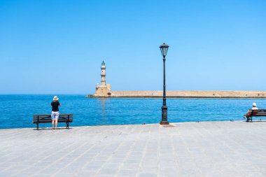 Chania Girit - 15 Ağustos 2024; Ortaçağ Venedik deniz feneri ve dalgakıran körfezi boyunca iki turist gezinti güvertesinde.