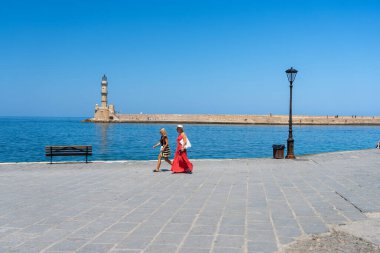 Chania Girit - 15 Ağustos 2024; Panama şapkalı ve uzun kırmızı elbiseli iki kadın arka planda deniz feneriyle gezinti güvertesinde yürüyor.