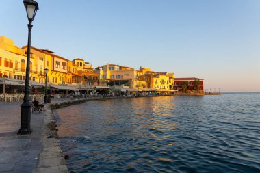 Chania Girit - 16 Ağustos 2024; Güneşin doğuşuyla birlikte gölgeler gezinti alanı ve kafelerin ön cephelerini kaplıyor ama körfezin etrafına parlıyor.