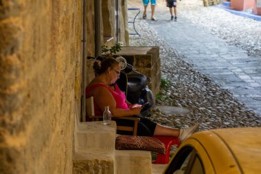 Rhodes, Yunanistan - 20 Ağustos 2024; Orta yaşlı bir kadın gece Rhodes Old Town 'da serin okuma arabasında kapısının önünde oturuyor.