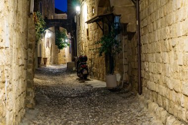 Rhodes, Yunanistan - 20 Ağustos 2024; Rhodes Eski Kasabası geceleyin loş ışıklandırılmış sokak sahnesindeki boş nehir taşlı sokak gölgesine park etmiş moped.