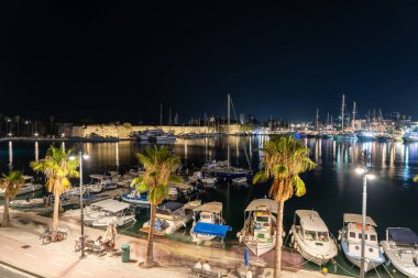 Limanda sallanan bulanık teknelerin olduğu gece sahnesi palmiye ağaçları ve yansıyan açık renklerle dolu..