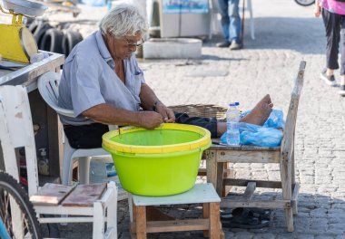Kos Yunanistan - 23 Ağustos 2024; Parlak sarı kovayla gölgede temizlik tezgahı ve tekne hazırlayan ve yem atan yaşlı adam.