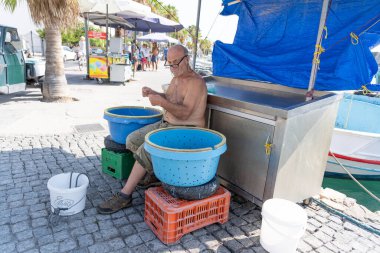 Kos Yunanistan - 23 Ağustos 2024; Geleneksel Avrupalı çıplak göğüslü, gözlüklü yaşlı adam temizlik tezgahının ve teknenin yanında oturmuş gölgeye hazırlanıyor ve yem atıyor.