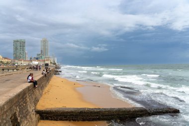 Colombo Sri Lanka - 2 Eylül 2024; Galle Face boyunca bulutlu gökyüzü altında insanlarla dolu deniz duvarı ve uzak ticari binalar.
