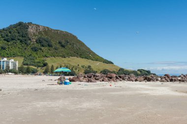 Piknik malzemeleri ve Maunganui Dağı arkaplanı üzerine ana plaj ve şemsiye.