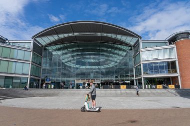 Norwich Birleşik Krallık - 7 Temmuz 2025; Norwich 'in elektrikli scooter kullanan bir adam ön plandan geçerken geçtiği zaman merkezdeki modern kamu binasına giden adımlar.