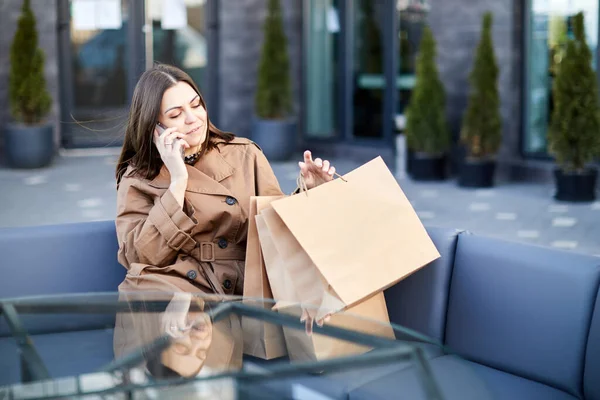木製のベンチに座っている若い女性の側のビュー新しいメッセージを介して携帯電話のスクロールを使用して購入と紙の袋 — ストック写真