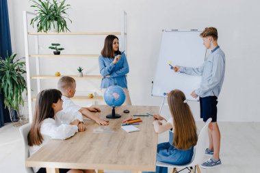 Coğrafya dersinde farklı yaşlardan bir öğretmen ve öğrenciler. Öğrenci tahtaya yakın durarak cevap verir ve hikayeyi anlatır.