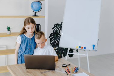 Ergen kız ve birinci sınıf öğrencisi çocuk laptopta birlikte ders çalışıyorlar sınıfta masa başında oturuyorlar. Genç kız ve birinci sınıf çocuğu okulda bilgisayar projesinde beraberler.
