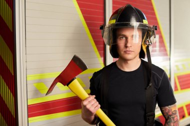 Photo of fireman wearing helmet with ax against fire engine. 