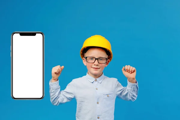 Cute boy construction worker in a yellow hard hat stands in an indoor studio on a blue background and shows satisfaction with the work done near huge mobile phone . Future construction worker