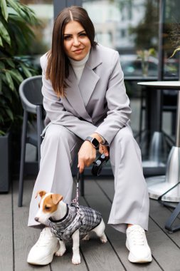 En sevdiği tüylü, eğitimli safkan Jack Russell Terrier köpeğiyle birlikte sonbahar günü kafede köpekler için takım elbise giymiş bir kadın oturuyor. Sadık en iyi arkadaşlar