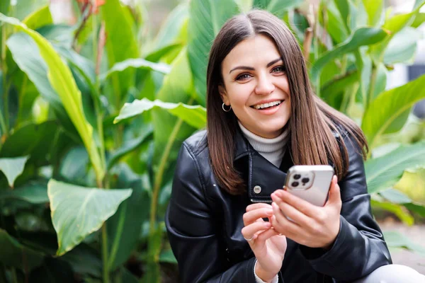 stock image A young pretty girl wearing a black leather jacket walks down the street in a park and uses her smartphone for work. The work of a freelancer and blogger