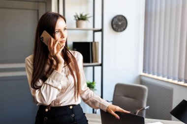 Business talk. Cheerful young beautiful woman talking on mobile phone and looking to the window while leaning on the table at her working place.