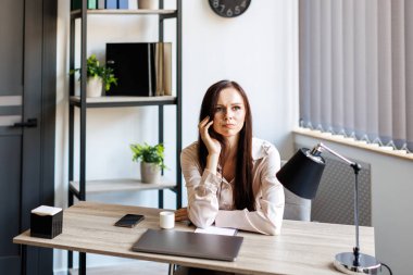 Bored female office employee sits at the desk in front of a laptop in the office, feels sad, a pensive young woman does not have inspiration for work, feels lack of sleep and fatigue. Overwork, working overtime and stress at work concept.