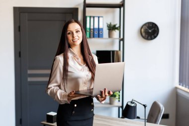 Adult successful female employee standing in modern office, hold a laptop in arms. Happy brunette-hair business lady in elegant stylish suit looks at camera and smiling