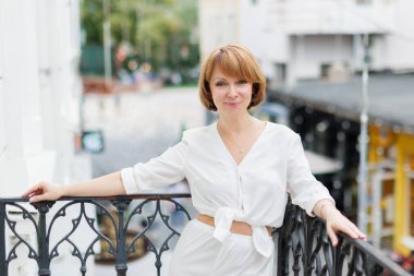 Beyaz takım elbiseli yetişkin bir kadın kasabanın balkonunda kameraya bakıyor.