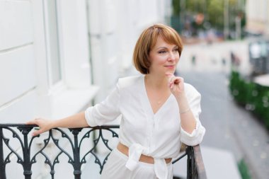 Beyaz takım elbiseli yetişkin bir kadın kasabanın balkonunda kameraya bakıyor.