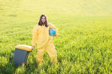 Sarı koruyucu takım elbiseli bir kadın yeşil bir tarlanın ortasında elinde bir küre tutarken diğer eliyle bir çöp tenekesi taşıyor..