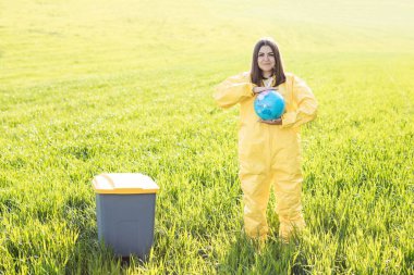 Sarı koruyucu takım elbiseli bir kadın yeşil bir tarlanın ortasında duruyor ve elinde bir küre tutuyor, çöp tenekesinin yanında. Gezegen bakımı, dünya kavramını sakla.