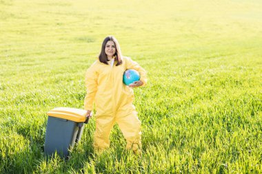 Sarı koruyucu takım elbiseli bir kadın yeşil bir tarlanın ortasında elinde bir küre tutarken diğer eliyle bir çöp tenekesi taşıyor..