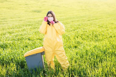 Sarı koruyucu takım elbiseli bir kadın yeşil bir tarlanın ortasında duruyor. Koruyucu bir maske takıyor, pembe eldivenler giyiyor..