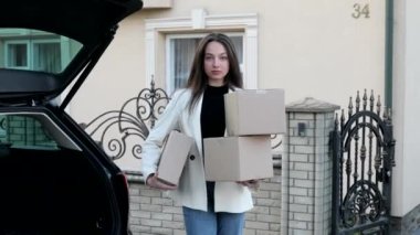 Young business woman picking up parcels from a car trunk, coming home by car. The girl carries boxes in her hands and they fall from her hands, she is upset