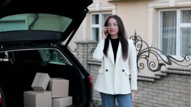 Young business woman standing at the car with parcels, coming home by car. The girl is standing and talking on mobile phone near the car. Concept of buying goods online and delivering them home