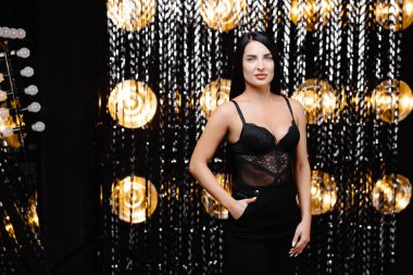 Young beautiful smiling brunette female in trendy evening clothes. Sexy carefree woman posing near shine wall in studio in a circle of light.Fashionable model with bright makeup.