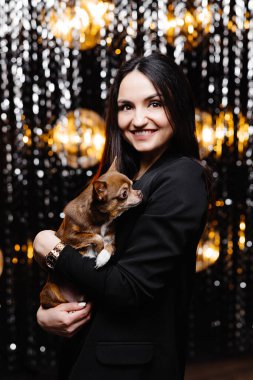 Young beautiful smiling brunette female in black jacket holding her litlle dog in her hands. Carefree woman posing near shiny tinsel wall in studio with dog