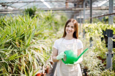 Genç bir kadın büyük bir seranın ortasında elinde bir su bidonuyla kameraya gülümsüyor.