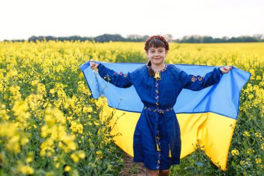 Ukrayna için dua et. Kolza tarlasında Ukrayna bayrağı taşıyan bir çocuk. Nakışlı gömlekli bir kız elinde Ukrayna bayrağıyla sahada koşuyor.