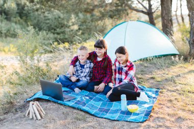 Ormanda dizüstü bilgisayar izleyen çocuklar. Erkekler ve kızlar bir çadır kampında dinleniyorlar. Çocuklar yazın çimlerin üzerinde ekose üzerine oturur. Yaz tatili, seyahat ve macera konsepti.