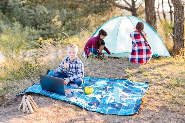 Lets go camping. Family camping. Reach destination place. Two girls pitch tent and one boy watching show on the laptop, sitting on the plaid. Hiking outdoor adventure.