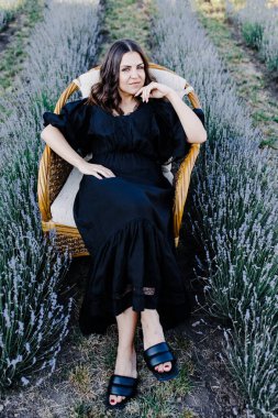 Attractive young woman in black dress and sitting in chair surrounded by lavender field.