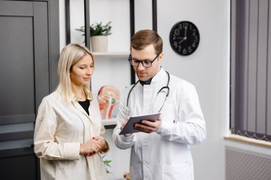 Happy man doctor showing tablet woman patient in clinic office interior. Health care, visit to family therapist, treatment of illness and medical help.