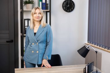 Masanın yanında duran mavi elbiseli çekici genç bayan. Ofis işyerinde poz veren genç profesyonel kadın..