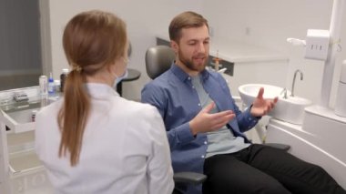 Blonde female dentist in dental office talking with male patient and preparing for treatment. Handsome bearded man in dentist chair looking at his doctor with smile