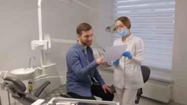 Young attractive man visiting dentist, sitting in dental chair at modern light clinic. Young woman dentist holding x ray image.