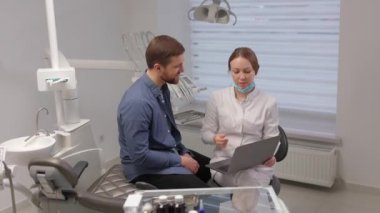 A handsome young man is talking to a female doctor at a dental appointment in a bright, beautiful office. The dentist explains to the patient and shows everything on the laptop