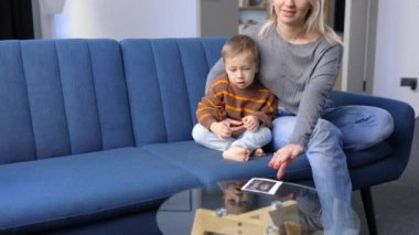 Young pregnant woman and her son sit on sofa at home and boy holds ultrasound scan in hand.They spend time together, enjoy communication feeling kicks of unborn baby