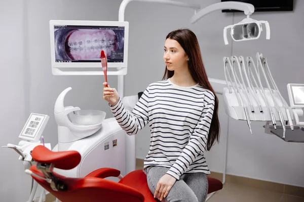 Happy caucasian female patient looking at mirror after dental treatment in clinic. Cheerful woman sitting in chair in dental cabinet and enjoying her beautiful smile.