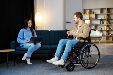 Tekerlekli sandalyedeki genç adam cep telefonunu elinde tutuyor ve karısı evde dizüstü bilgisayar kullanıyor..